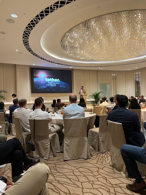 A conference room scene showing a presentation about 'tether' (described as 'A global ecosystem for innovation') with an audience seated at round tables beneath an ornate crystal chandelier.
