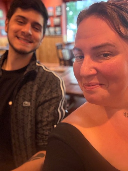 A selfie-style photo of a smiling young man and a woman with short dark hair sitting together in what appears to be a restaurant or bar.