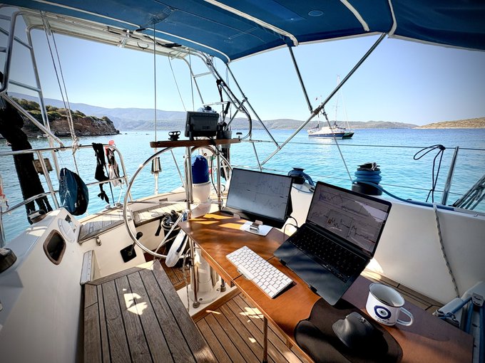 The cockpit of a sailboat serves as a scenic mobile office, equipped with a laptop, external monitor, keyboard, and coffee mug overlooking calm turquoise waters and anchored yachts. The post humorously contrasts the typical Monday work grind with the luxury of working remotely aboard a sailboat in a picturesque harbor.