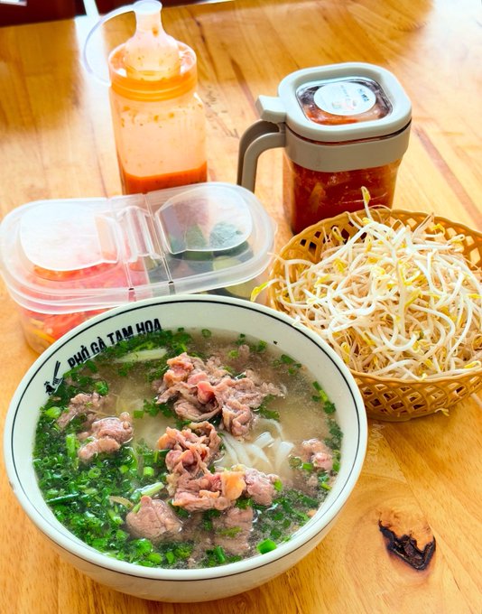 A bowl of Vietnamese pho labeled 'PHỐ GÀ TÂM HÒA' containing beef, noodles, and green onions, served with a basket of bean sprouts and two bottles of chili sauce on a wooden table.