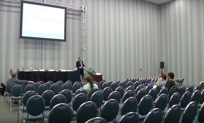 A speaker in a suit addresses a small audience in a conference room with rows of empty chairs and a projection screen displaying a presentation.