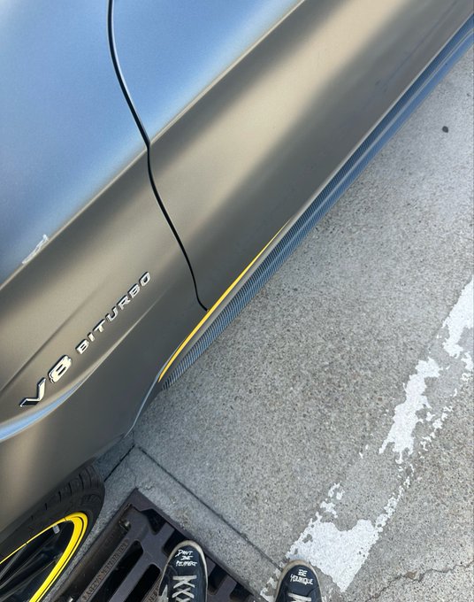 Close-up of a matte gold Mercedes V8 Biturbo vehicle with a yellow accent stripe along the side skirt, parked on asphalt next to someone wearing Travis Scott sneakers.