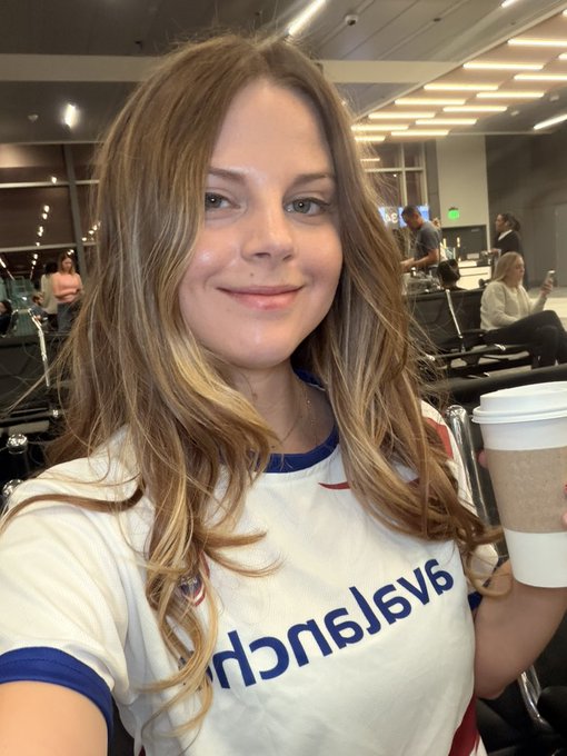 A young woman with long brown hair wearing a white Chelsea F.C. jersey takes a selfie in what appears to be an airport while holding a coffee cup.