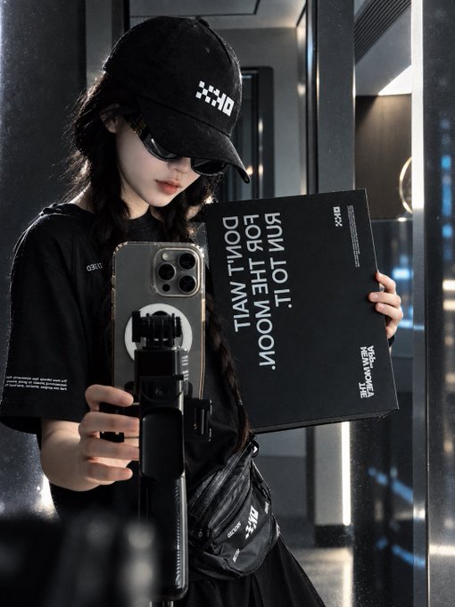 A woman wearing sunglasses, a black cap with a white logo, and a black t-shirt takes a mirror selfie with a phone gimbal while holding a black book titled 'KIM JOLI: FOR THE MOON, DON'T WAIT.'