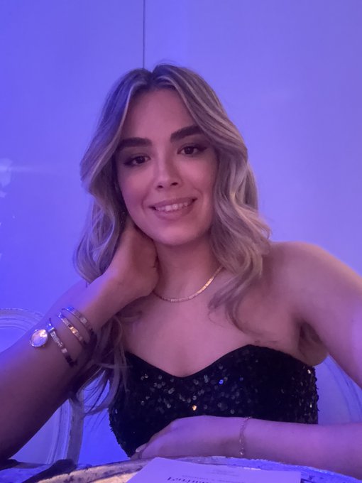 A young woman with wavy blonde hair smiling at the camera, wearing a black sequined strapless top and gold jewelry, seated against a vibrant purple-lit background.