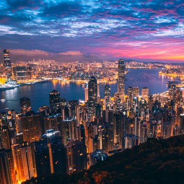 The image shows a stunning aerial view of Hong Kong's skyline at sunset, with the city's illuminated skyscrapers reflecting off the harbor under a vibrant sky of pink and purple hues. This visual captures the financial hub at the heart of the news about Hong Kong's plans to license crypto trading, custody, advisory, and management services within the year.
