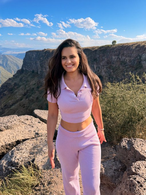 A woman with long brown hair wearing a light pink two-piece outfit stands smiling at the camera with a dramatic canyon landscape in the background.