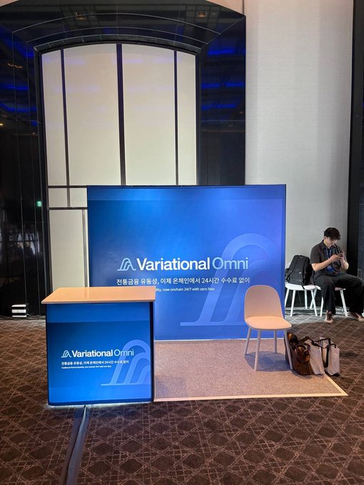 A blue promotional booth for "Variational Omni" featuring Korean text about special perks for attendees, set up in a convention hall with a person sitting at a nearby table.