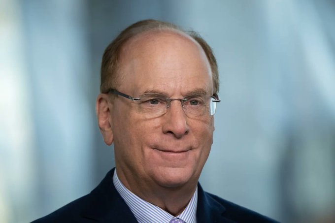 A professional headshot of an older man with glasses, wearing a dark suit, striped shirt, and tie, smiling slightly against a blurred blue background.