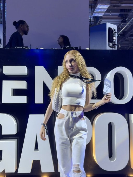 A woman with curly blonde hair wearing a white crop top and pants stands on a stage at an 'ENERGY GANG' event, with a DJ booth visible in the background.