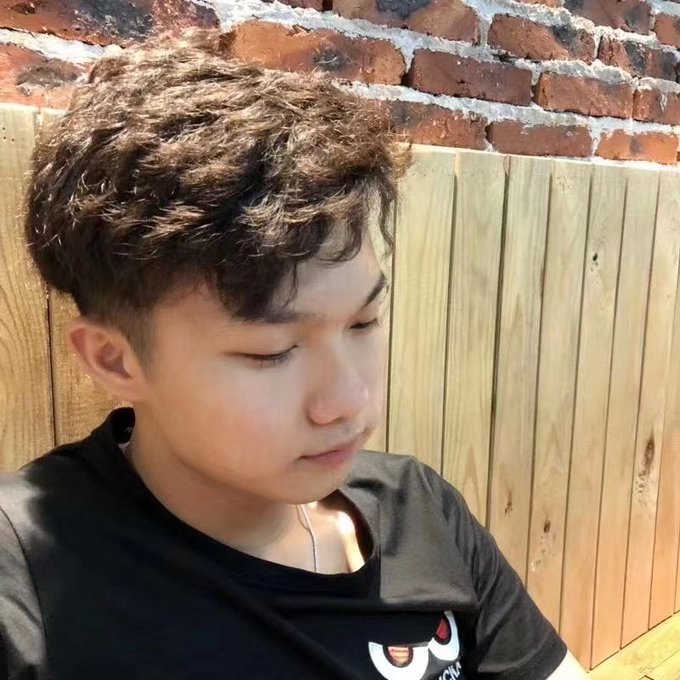 A young man with curly brown hair and a black Champion t-shirt looks down pensively in front of a brick wall and wooden fence. The image visually conveys the post's theme of self-deprecating frustration, as his contemplative expression reinforces the caption's complaint about having looks but no monetary value—echoing the sentiment that his handsomeness can't be converted into cash or crypto gains despite his airdrop efforts.