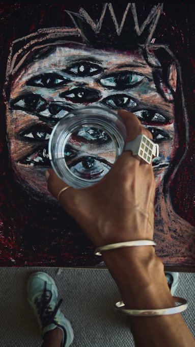 A close-up of a hand wearing silver rings and bracelets holding a glass of water, with a textured painting featuring multiple eyes and a crown visible in the background.