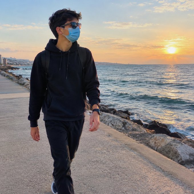 A person wearing a black hoodie, sunglasses, and a blue face mask walking along a coastal path at sunset.