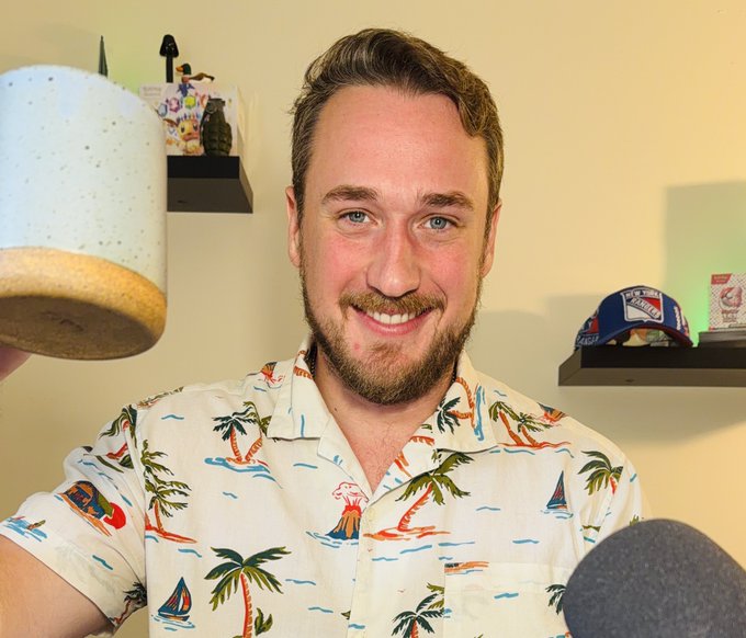 A smiling bearded man in a tropical-print Hawaiian shirt holds up a white speckled ceramic mug, with a microphone visible in the foreground and shelves displaying decorative items behind him.