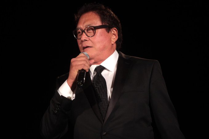 The image features an older man wearing glasses and a suit standing on stage with his hand raised to his mouth, possibly speaking into a microphone or delivering a speech. The background of the image is black, which contrasts with the white text that reads "LATEST: Rich Dad Poor Dad author Robert Kiyosaki warns that most investors are de-worsified, not diversified." This statement emphasizes the importance of investing in diverse assets to avoid being de-worsified by market crashes.