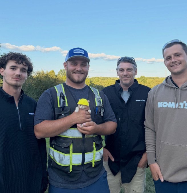 Four men wearing Komatsu branded clothing and safety vests pose outdoors with one man in the center holding a small hamster wearing a yellow hard hat.