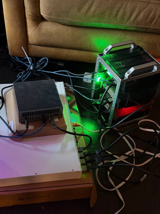 An ASUS router and a "MIKROTIK" device are visible, surrounded by a tangled mess of cables, with a green LED illuminating the area. The image accompanies a post about "4 x DGX = GLM 5.2 20Tok/s," though the specific DGX hardware is not clearly visible in the frame.
