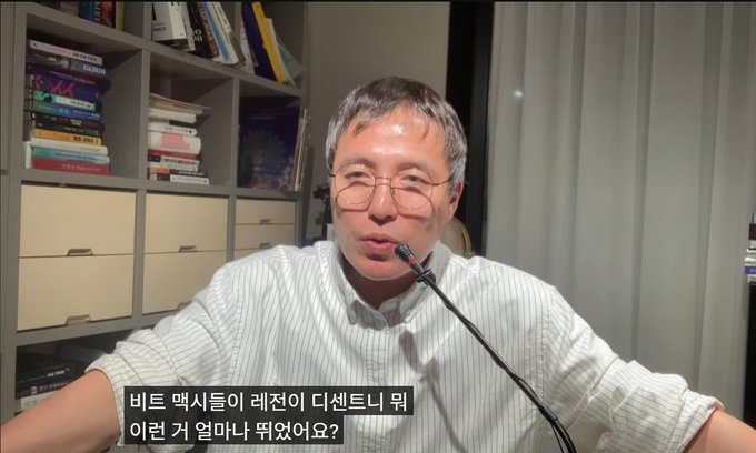 The image shows a middle-aged man with glasses, wearing a white striped button-up shirt, speaking into a gooseneck microphone in what appears to be a home office setting with a bookshelf in the background. Korean subtitles at the bottom of the image read "비트 맥시즘이 레저니 디센터니 뭐 이런 거 얼마나 띄웠어요?" which translates to the portion of the quote about how much Bitmaxis hyped up Ledger and Decent, matching the commentary about hardware wallet promotion.