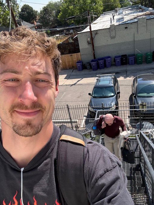 A young man with brown hair takes a selfie outdoors, smiling, while an older man walks down metal stairs behind him carrying bags, with parked cars and trash bins visible in the background.