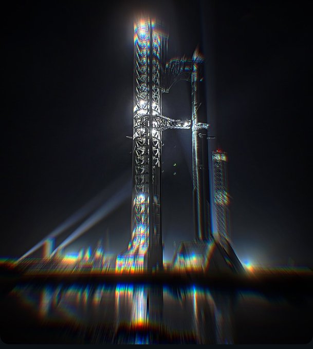 A towering, intricately detailed structure resembling SpaceX's Starship launch tower stands against a dark background, with the image stylized with rainbow chromatic aberration and reflected on a water-like surface below, evoking the surreal, hallucinatory vibe of the post's "trippy" descriptor.
