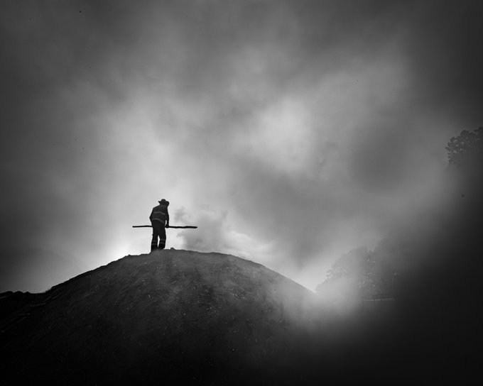 A black-and-white silhouette of a figure wearing a wide-brimmed hat and holding a long staff stands atop a hill against a cloudy, misty sky.