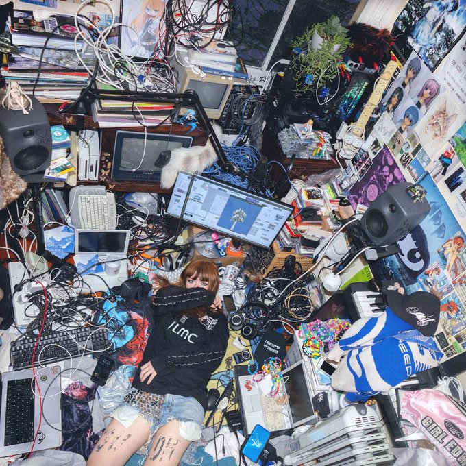 A top-down photograph of a person lying on the floor in a chaotic room filled with tangled wires, monitors, audio equipment, books, plush toys, and posters.