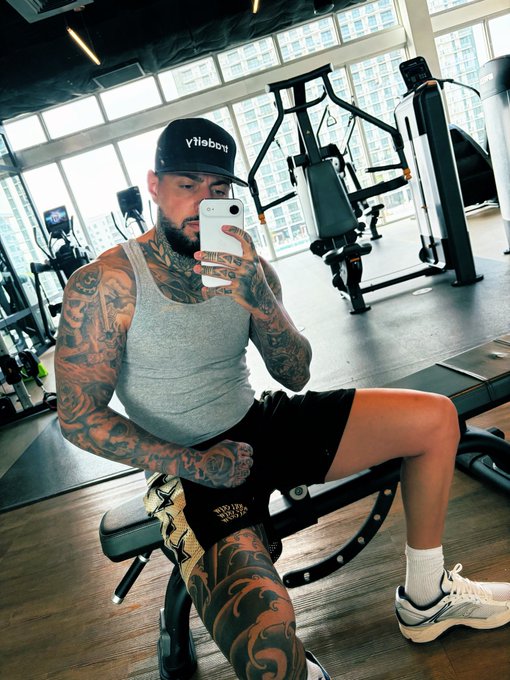A heavily tattooed man takes a mirror selfie in a gym while seated on a weight bench, wearing a "YaBoi" cap, gray tank top, and black shorts, with the message emphasizing disciplined training on Sundays when others rest as a metaphor for outworking competitors in the markets.