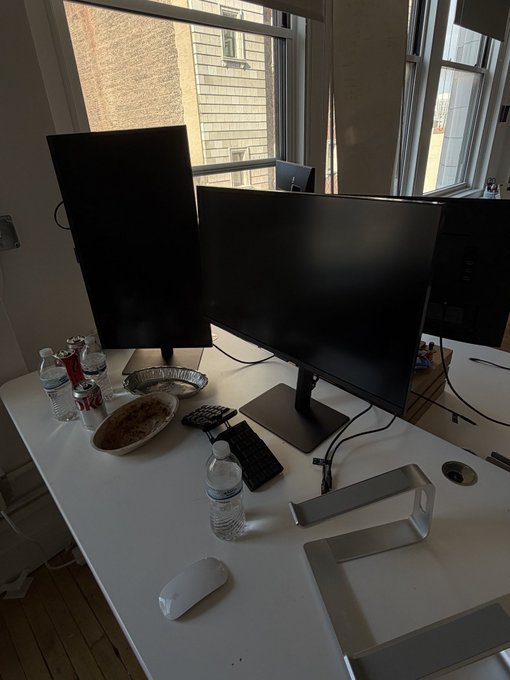 A home office desk setup featuring a vertical and horizontal monitor, a keyboard, mouse, water bottles, a can of Coke, a bowl of food, and a foil dish, all near a window.