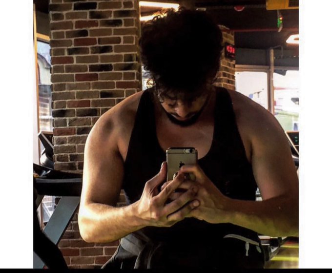 A muscular man in a black tank top sits in what appears to be a gym, intently focused on his smartphone as he types. The image complements the "Gm Web3 Folks" morning greeting, capturing the casual "good morning" check-in vibe common in the crypto and Web3 community, where members often start their day by scrolling through messages and market updates on their phones.