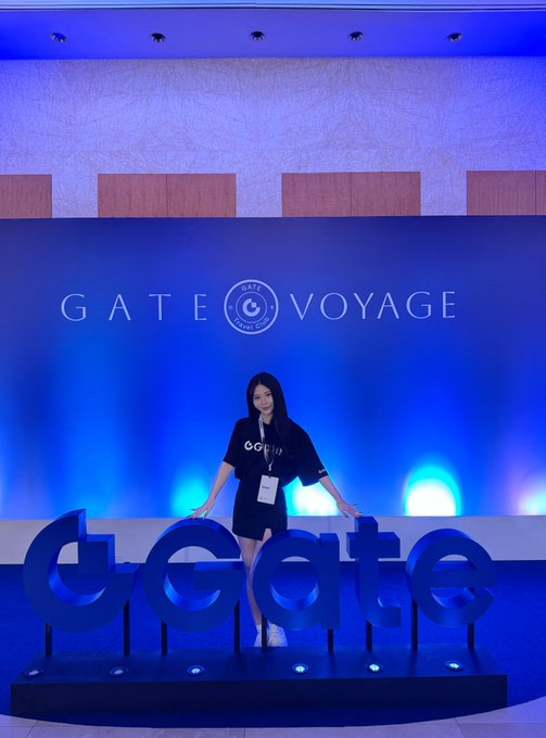 A woman wearing a black outfit and lanyard stands between large blue 'dGate' letters in front of a blue backdrop featuring the 'GATE VOYAGE' logo.