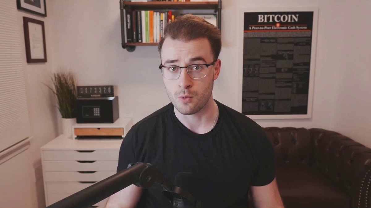 The image shows a man wearing glasses sitting behind a microphone with his hands on his knees, giving him an intense look as if he's about to speak into the microphone or record something important. The background features a bookshelf filled with various books, suggesting that this scene takes place in a room dedicated to reading and learning. A poster is also visible in the image, which appears to be related to Bitcoin.