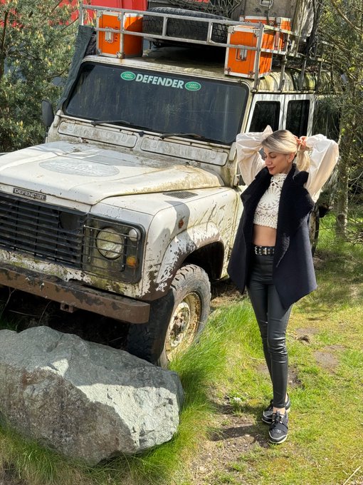 A woman with blonde pigtails, wearing a black fur vest, white lace top, and black leather pants, stands posing in front of a mud-splattered white Land Rover Defender that has orange cases strapped to its roof rack, with "DEFENDER" visible on the windshield.