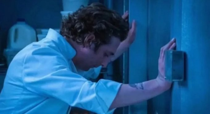 A man in a white shirt leans his head against a metallic door or wall in a dimly lit, blue-toned room, appearing distressed.