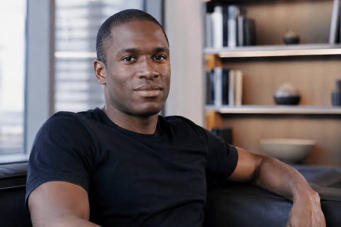 A portrait of a man in a black t-shirt sitting on a leather couch, smiling at the camera with a bookshelf and window visible in the background.