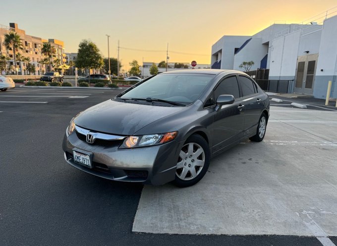 A gray Honda Civic sedan is parked on a curb during sunset, with buildings and palm trees in the background.