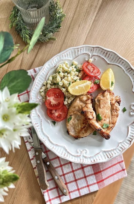 A plate of grilled chicken thighs served with pasta, sliced tomatoes, and lemon wedges, accompanied by a fork and knife on a red-checkered napkin.