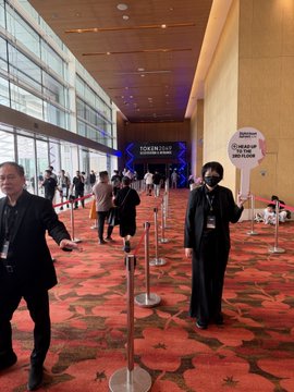 A convention or event space with people wearing masks, featuring signs that read "TOKEN 2049" and "CAPITALISM IS THE REAL CRIME!".