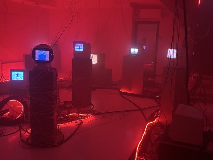 An art installation featuring multiple old CRT televisions and monitors displaying footage, bathed in red lighting with tangled cables throughout the room.