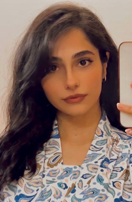 A woman with long dark hair takes a mirror selfie wearing a white blouse with a blue and brown paisley pattern.