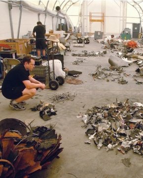A photograph showing two people examining scattered debris and scrap metal inside a workshop structure.