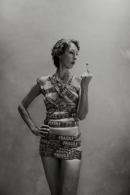 A black-and-white artistic photograph of a woman wearing a dress made entirely of 'FRAGILE' tape, posing with her middle finger raised.