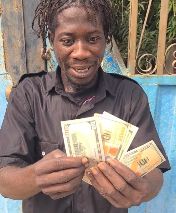 A smiling man with dreadlocks wearing a black shirt, holding and displaying several US dollar bills.