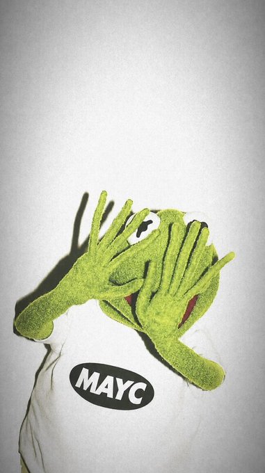 Kermit the Frog puppet lies face-down on a white cloth featuring a "MAYC" logo, capturing the iconic "HMM" moment of contemplative thought often associated with the character.