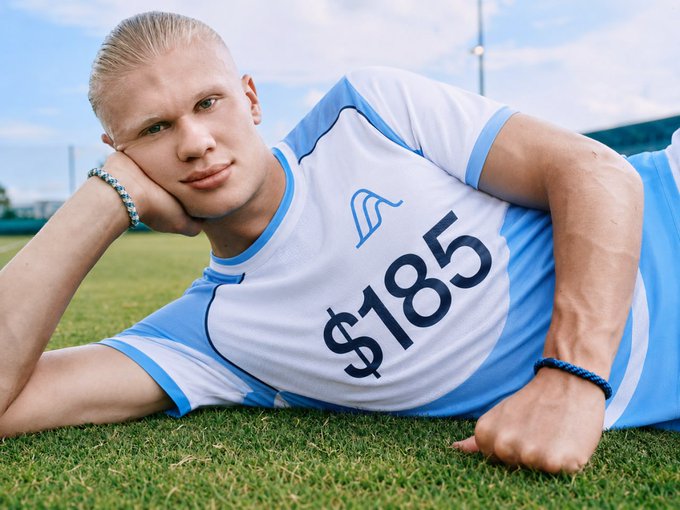 A young man with blonde hair lies on a grass field wearing a blue and white sports jersey featuring a wave logo and the text '$185'.