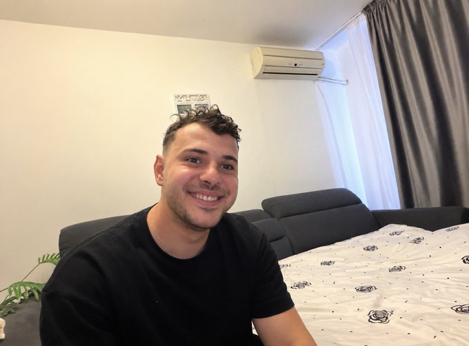A man in a black t-shirt smiles at the camera while sitting in a bedroom with a bed, gray curtains, and an air conditioner visible behind him. The post details his July achievements, and the image likely represents him relaxing in his personal space after accomplishing milestones like finishing his master's degree and earning money from content creation.
