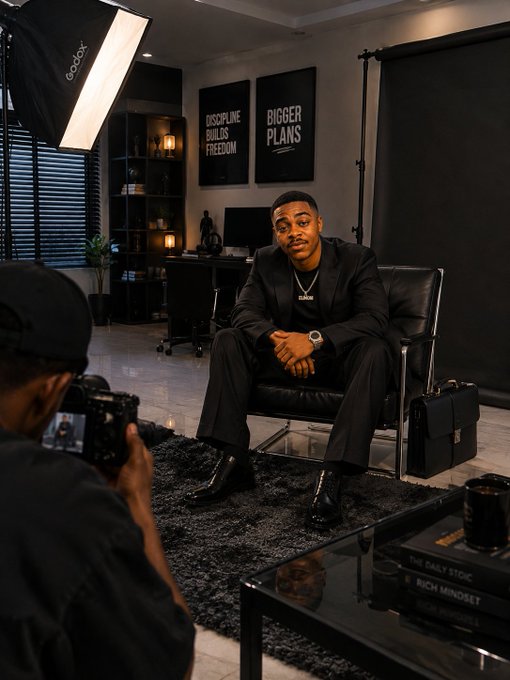 A man in a dark suit and gold chain sits in a modern office chair being photographed by someone with a camera in the foreground; framed posters behind him read 'DISCIPLINE BUILDS FREEDOM' and 'BIGGER PLANS', and books on the table include 'THE DAILY STOIC' and 'RICH MINDSET'.