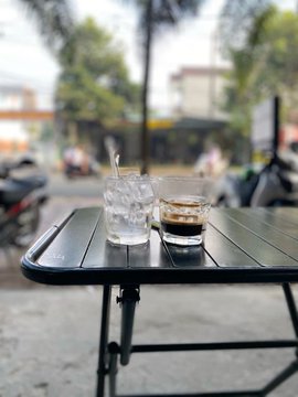 Two glasses containing iced and clear beverages sit on a black slatted outdoor table, with a blurred street scene visible in the background.