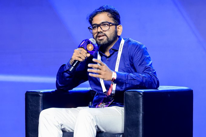 A man wearing glasses and a blue shirt speaks into a microphone while seated in a black chair on a stage with blue lighting, wearing a lanyard with a pass.