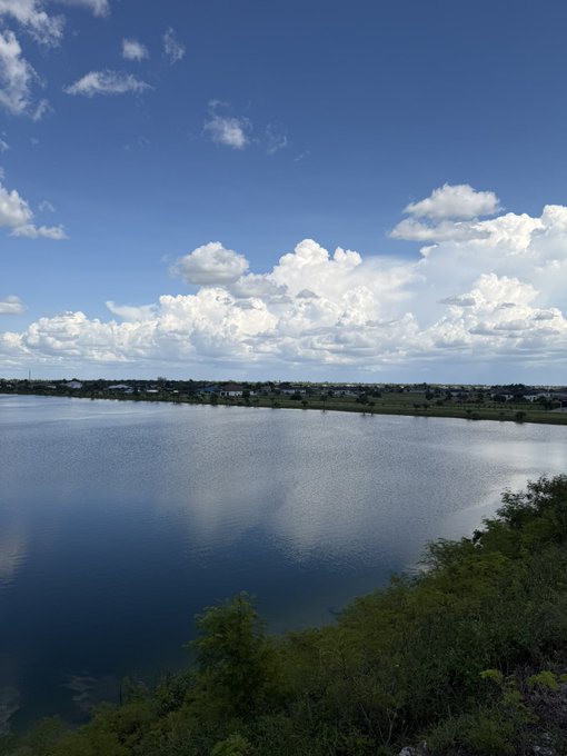A scenic landscape photo of a calm lake reflecting a partly cloudy blue sky, with green vegetation in the foreground and distant shoreline.