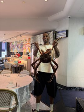 A man poses enthusiastically holding up a massive crab in a brightly decorated restaurant, celebrating Ansem's lavish $1400 buffet splurge in Singapore—a far cry from the ramen-or-soup budget he had once agonized over.